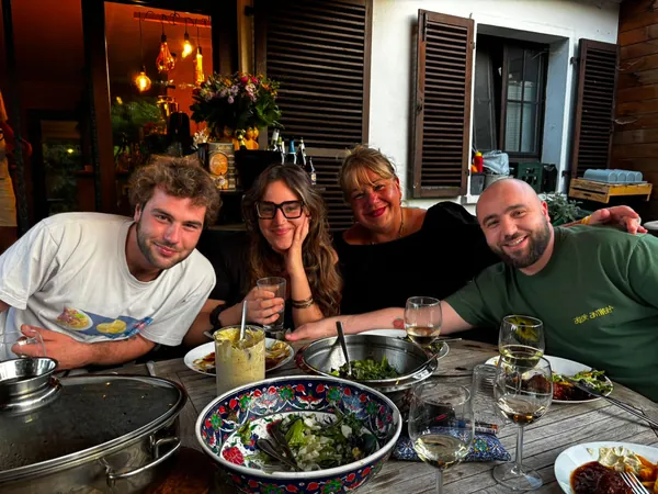 Quatre personnes de la maison attablées dehors, grands plats à partager et verres de vin blanc